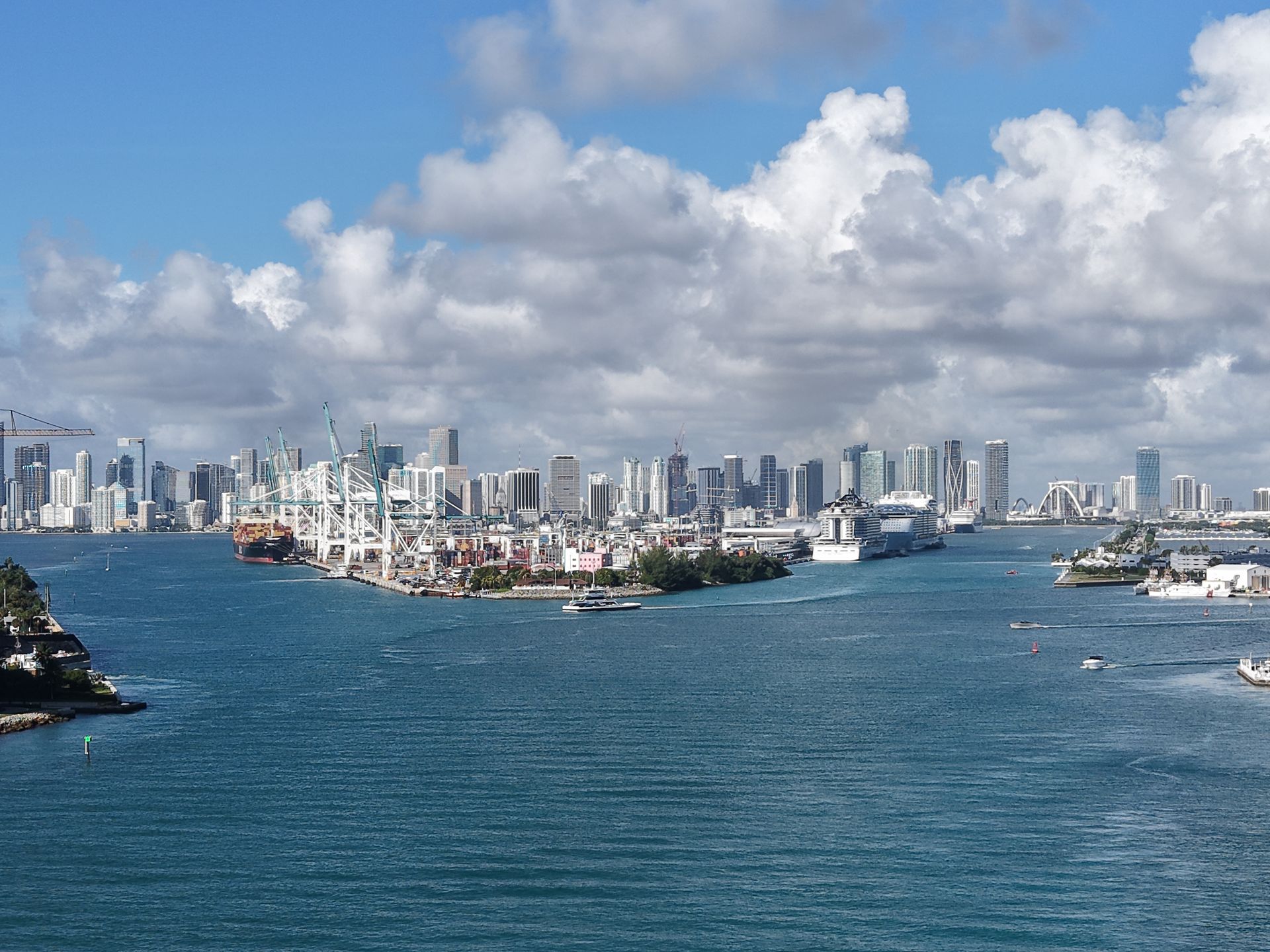 A view of the Miami skyline across the water, featuring a bustling port with docked cruise ships under a cloudy sky.