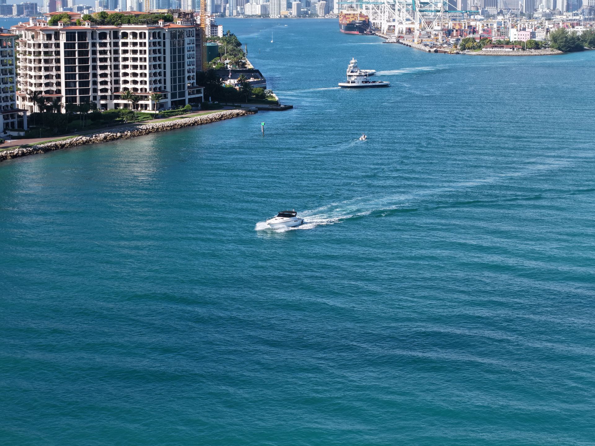 A white boat travels through a blue channel near a large apartment building and a city skyline under a clear blue sky.