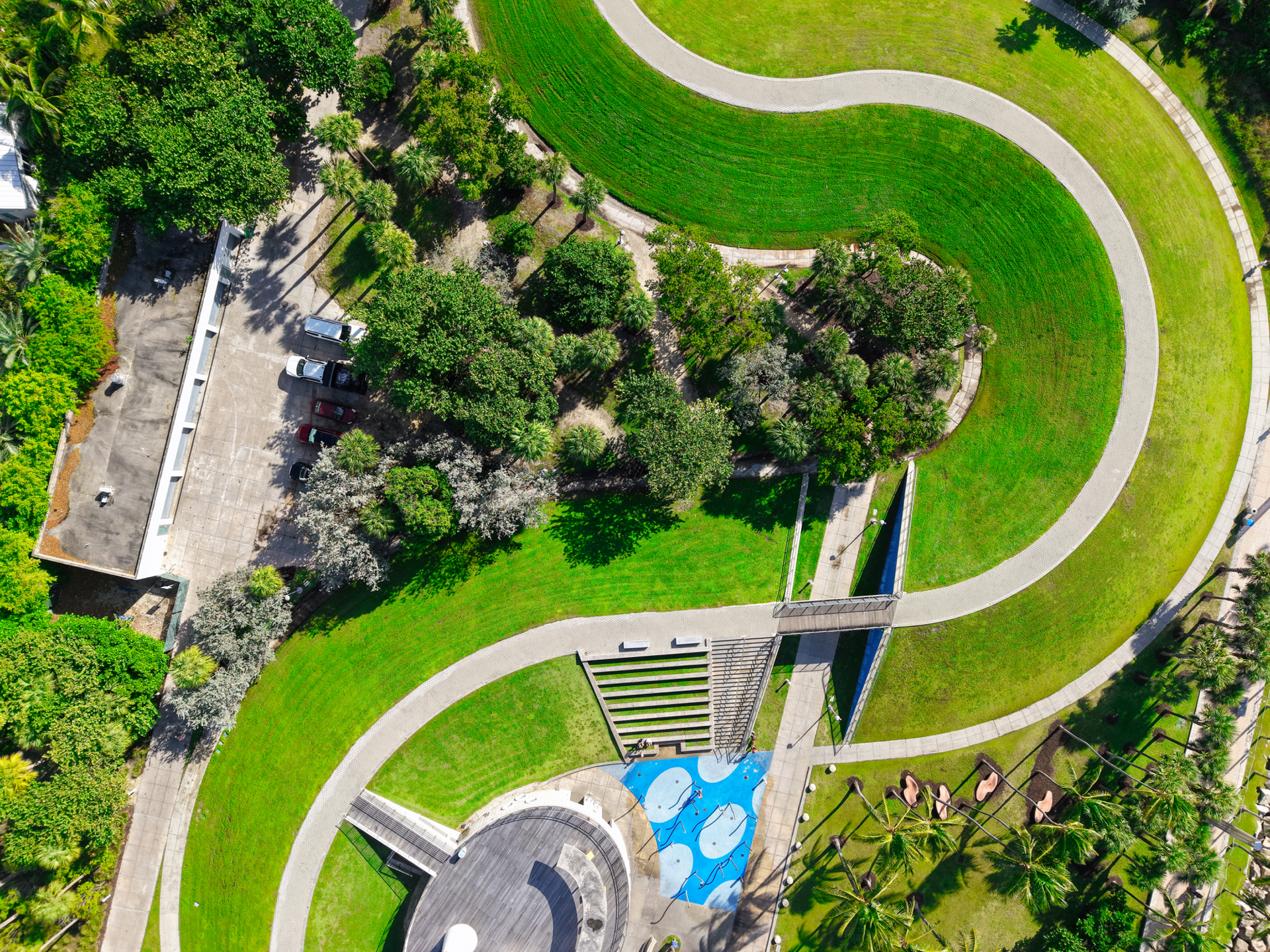 Aerial view of a park with curved walkways, a green lawn, a small parking area, and a blue playground section.