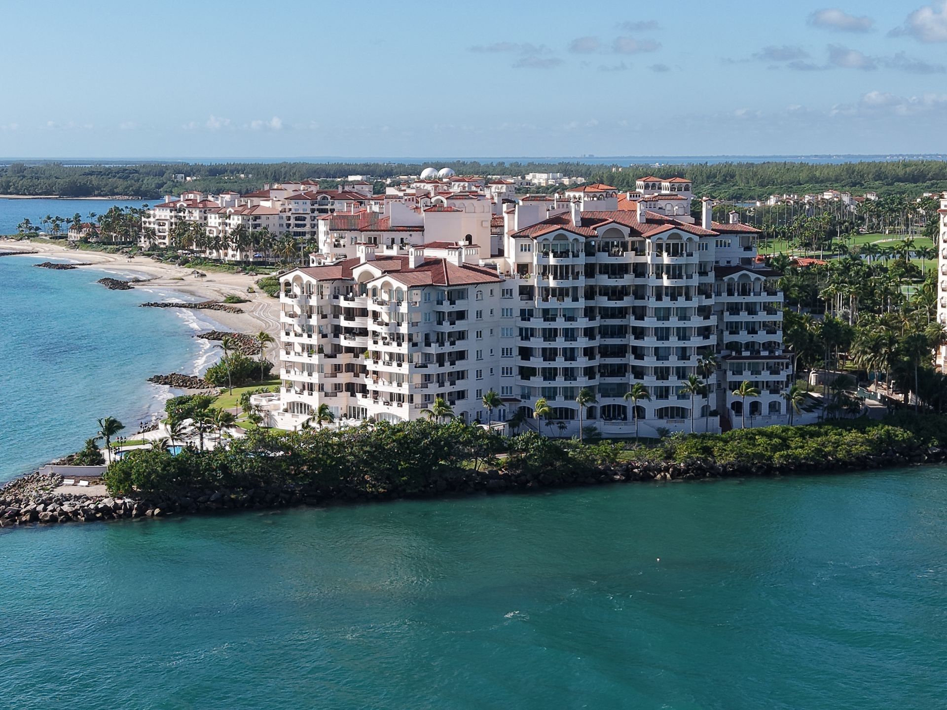A white, multi-story coastal apartment complex sits on a point of land surrounded by turquoise water and green foliage.