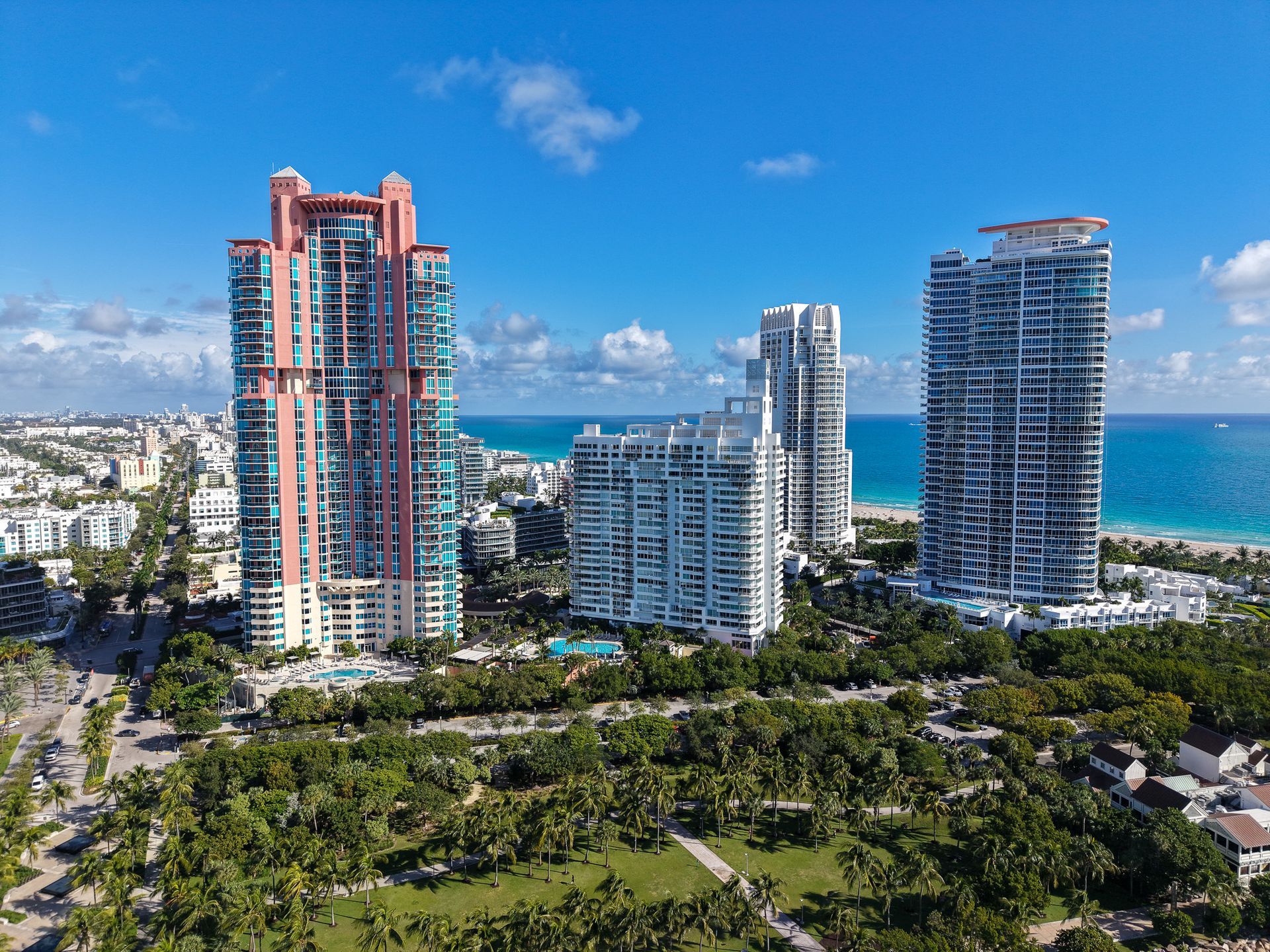 An aerial view of three high-rise apartment towers surrounded by lush greenery along a bright, turquoise coastline.