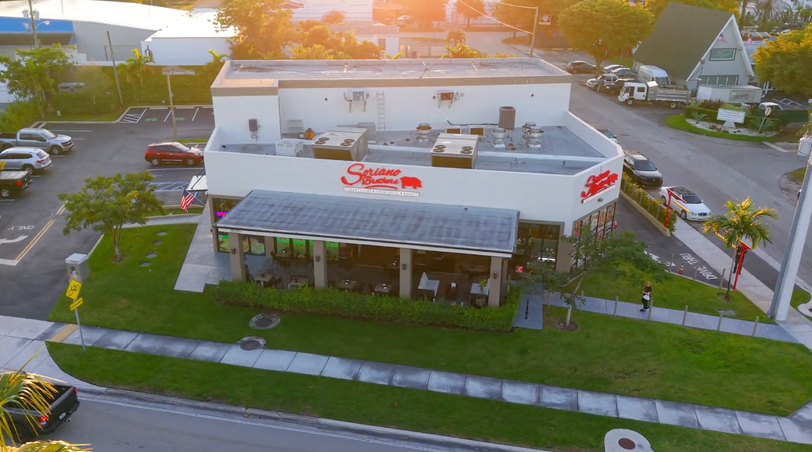 Restaurant building with outdoor seating, red sign, and surrounding green lawn.