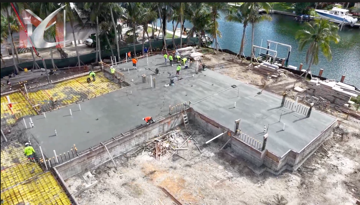 Construction site with workers pouring concrete foundation near water, palm trees.