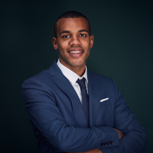 Man in blue suit, arms crossed, smiling, set against a dark teal background.