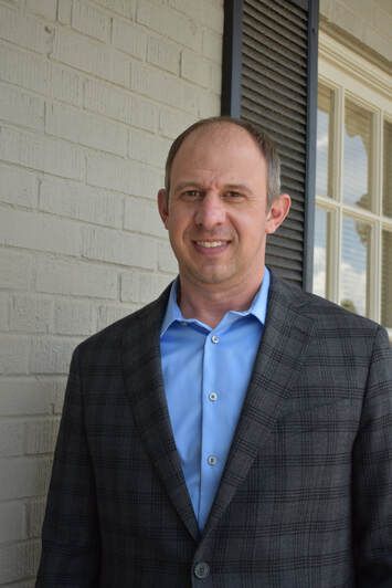 A man in a suit and blue shirt is standing in front of a brick building.