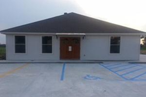 Small white building with a dark roof and brown door, viewed from a parking lot with blue handicap lines.