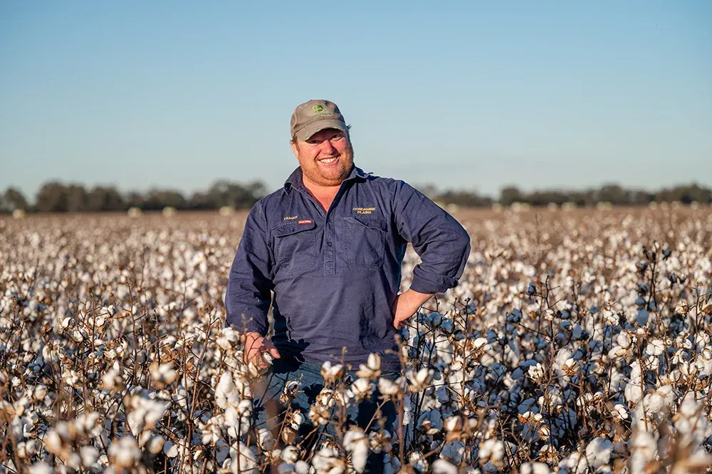 Farmer standing in a cotton field under a clear blue sky, wearing a navy shirt and cap.