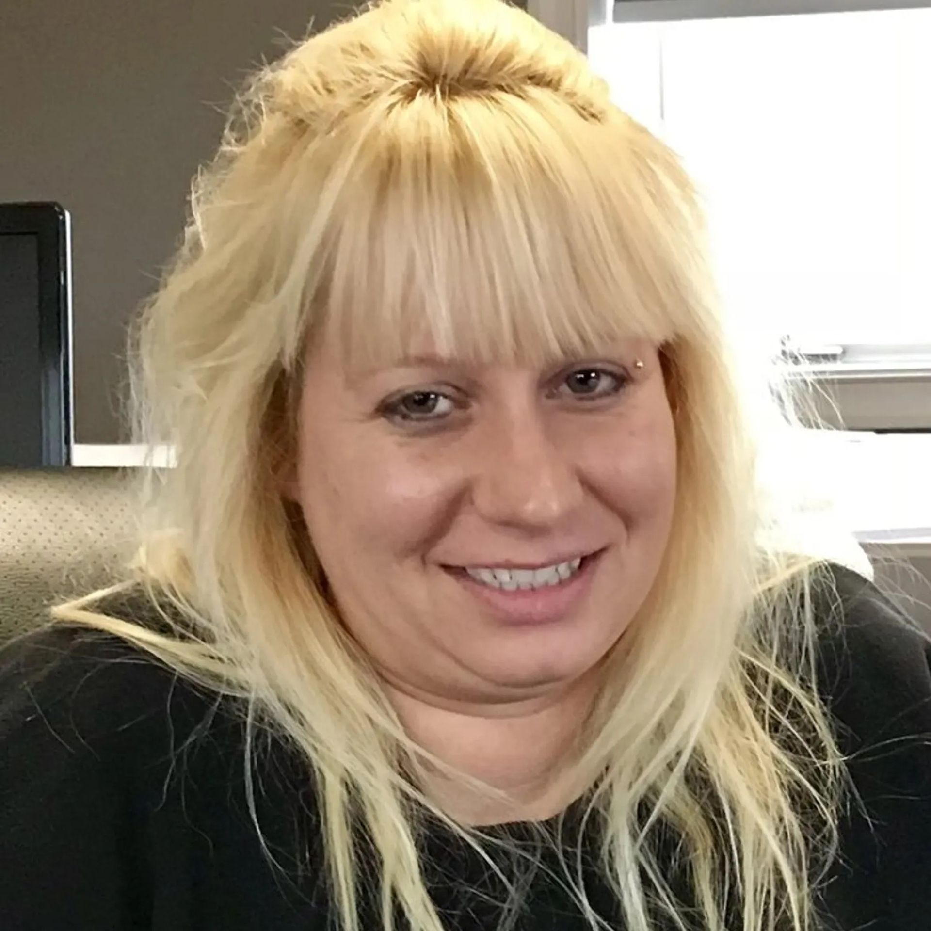 Smiling blonde woman in a black top indoors near a bright window