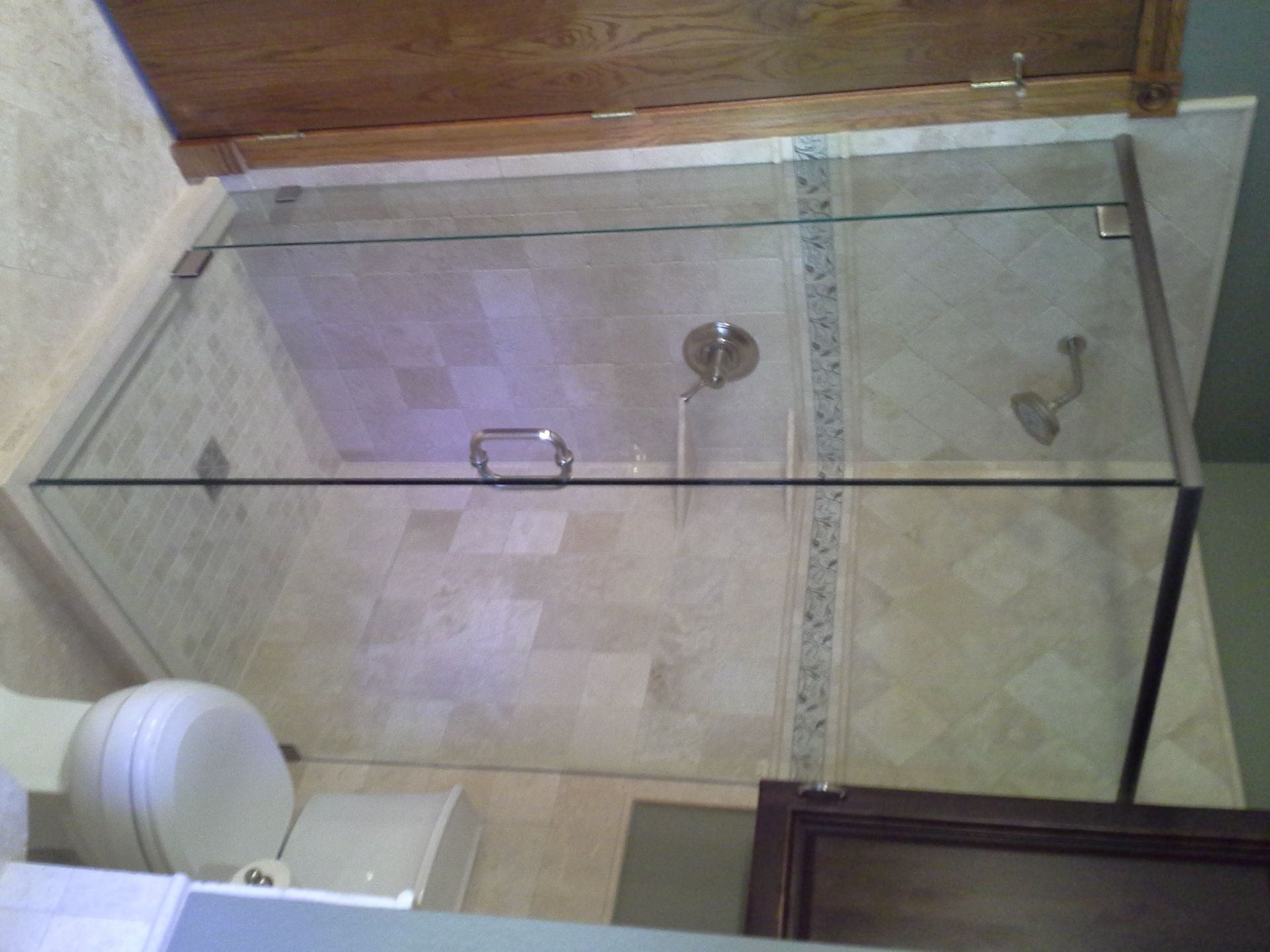 Glass shower enclosure in a tiled bathroom with beige walls and a white toilet visible outside.