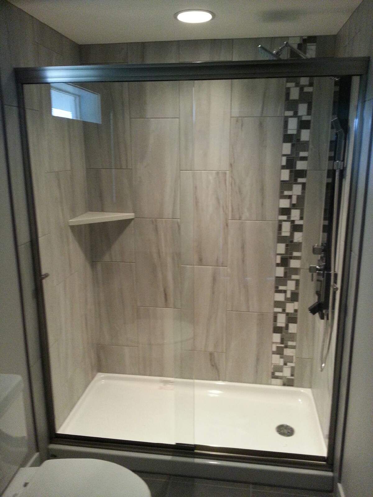 Accessible walk-in shower with glass door, gray tile walls, shelf, and white shower base.