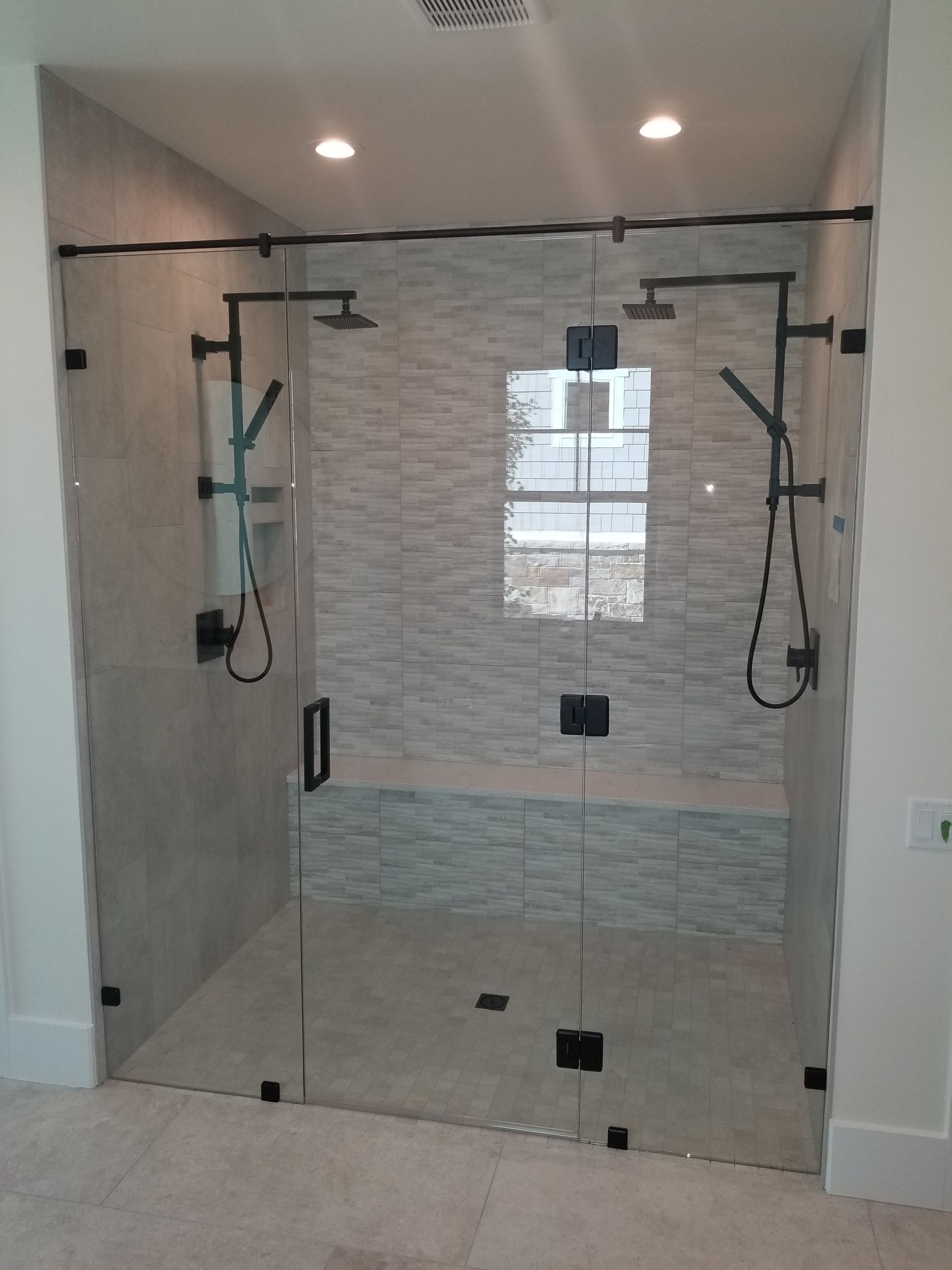 Glass shower enclosure with two black showerheads and a window reflection in a tiled bathroom
