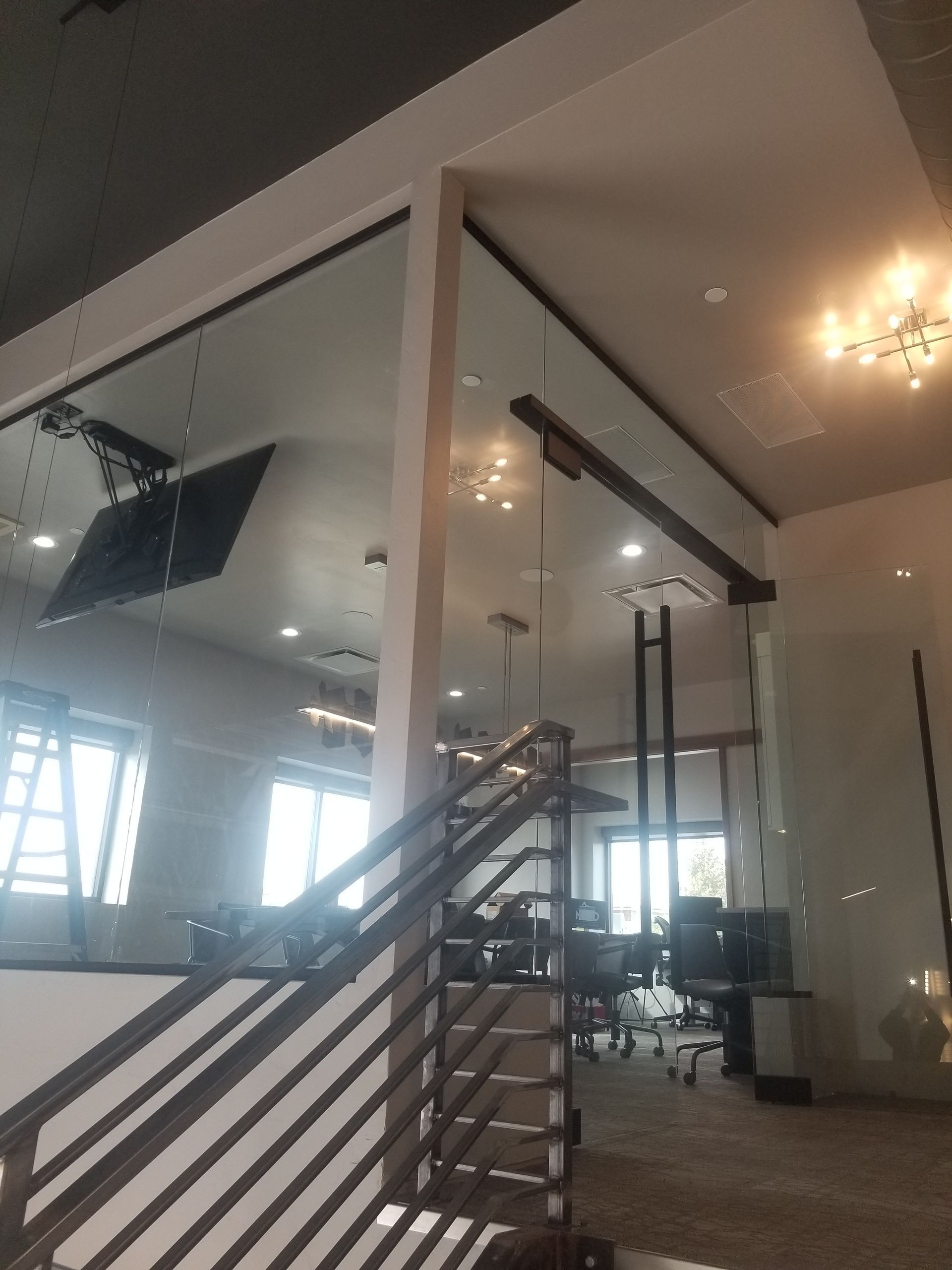 Modern living room with staircase, glass partition, and ceiling lights; bright windows reflect in mirrors