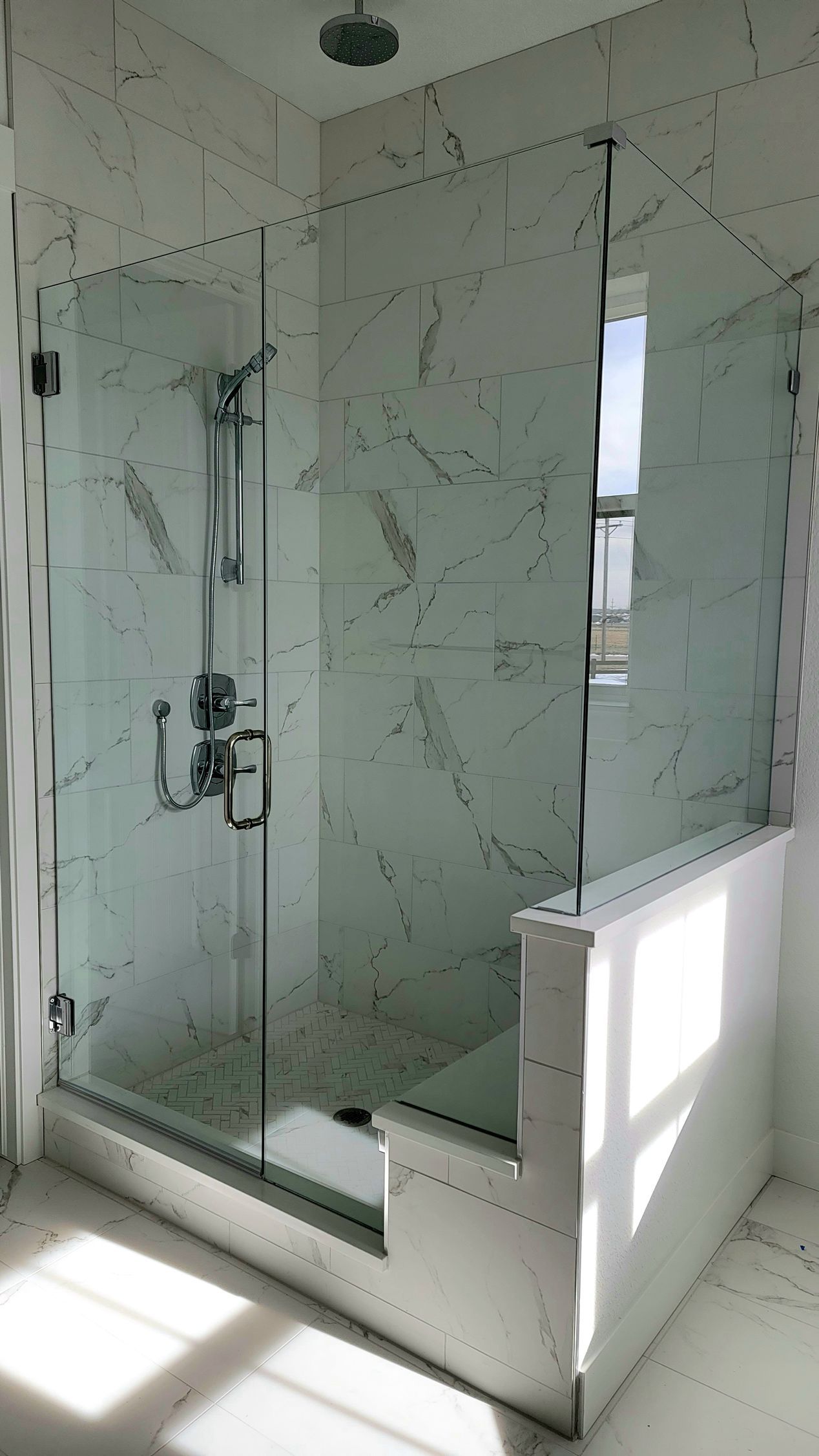 Modern tiled walk-in shower with glass enclosure, rain showerhead, and bright sunlight on the floor