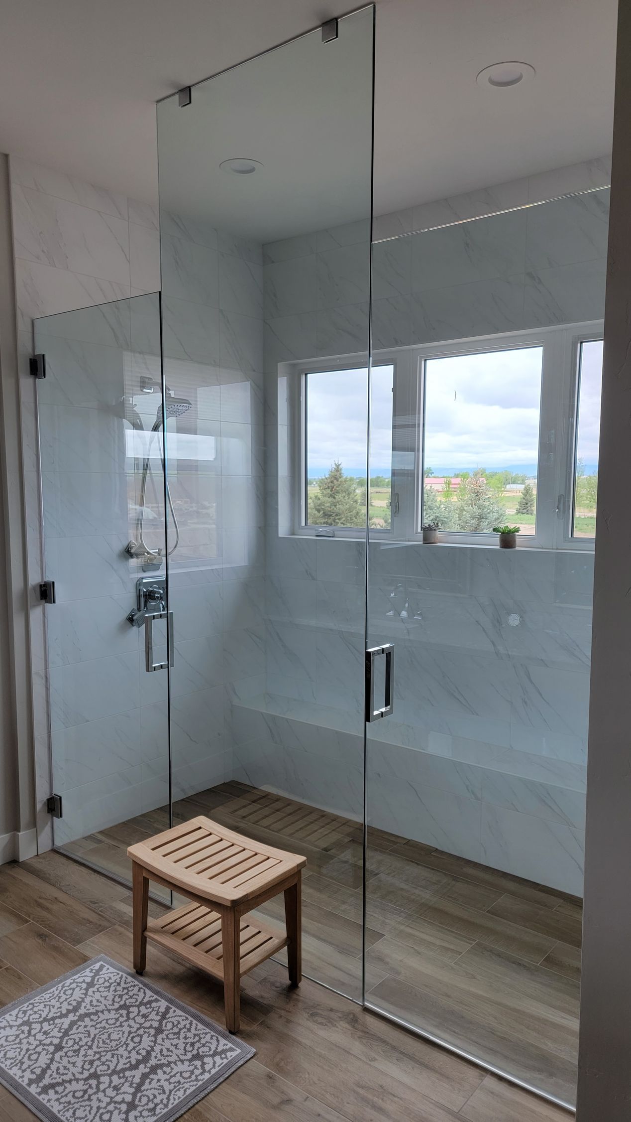Glass shower enclosure with chrome handles in a bright bathroom, with a small wooden stool outside.