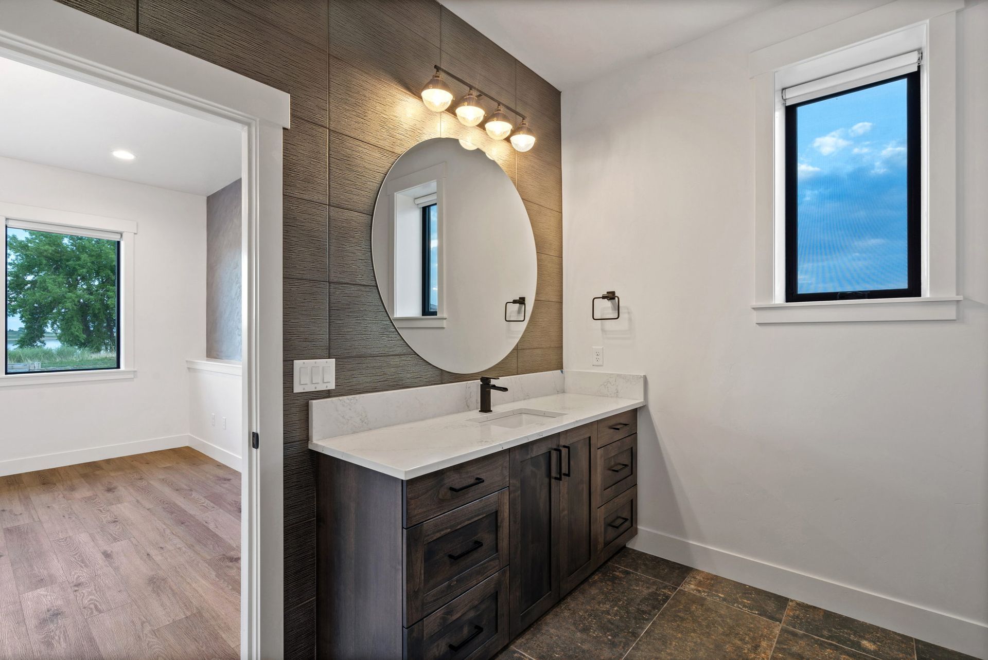 Modern bathroom vanity with round mirror, wall lights, and small window showing blue sky