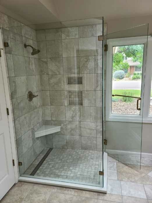 Walk-in tiled shower with glass door, bench, and small window overlooking a yard