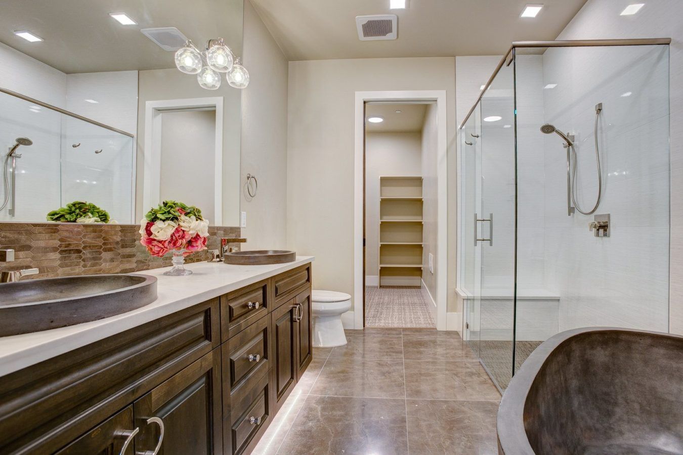 Modern bathroom with dark wood vanity, large mirror, glass shower, and tiled floor, viewed toward a walk-in closet