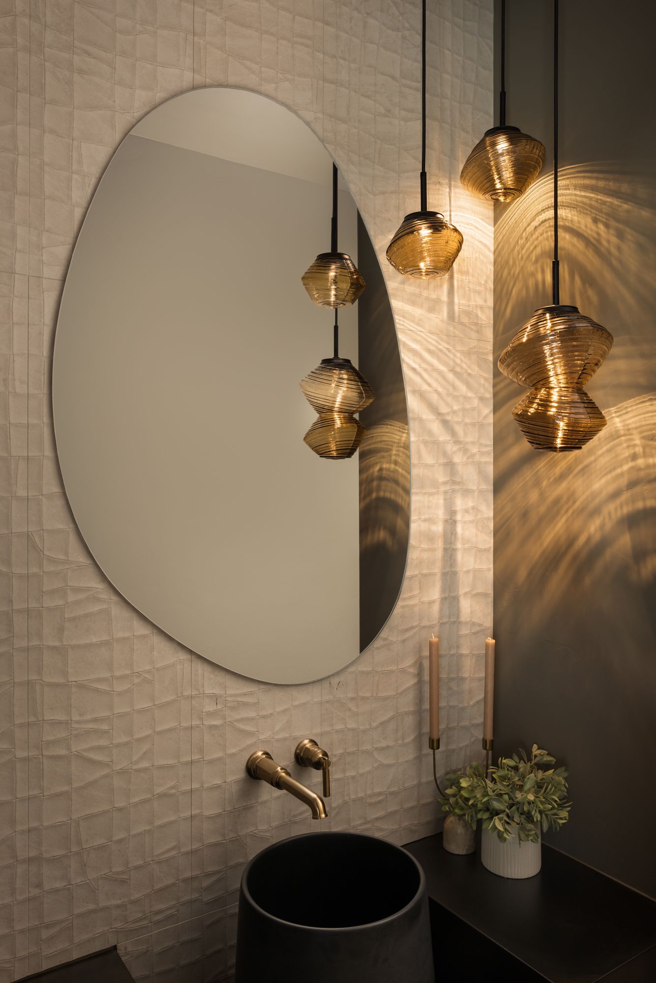Modern bathroom vanity with oval mirror, black sink, gold pendant lights, and small potted plant.