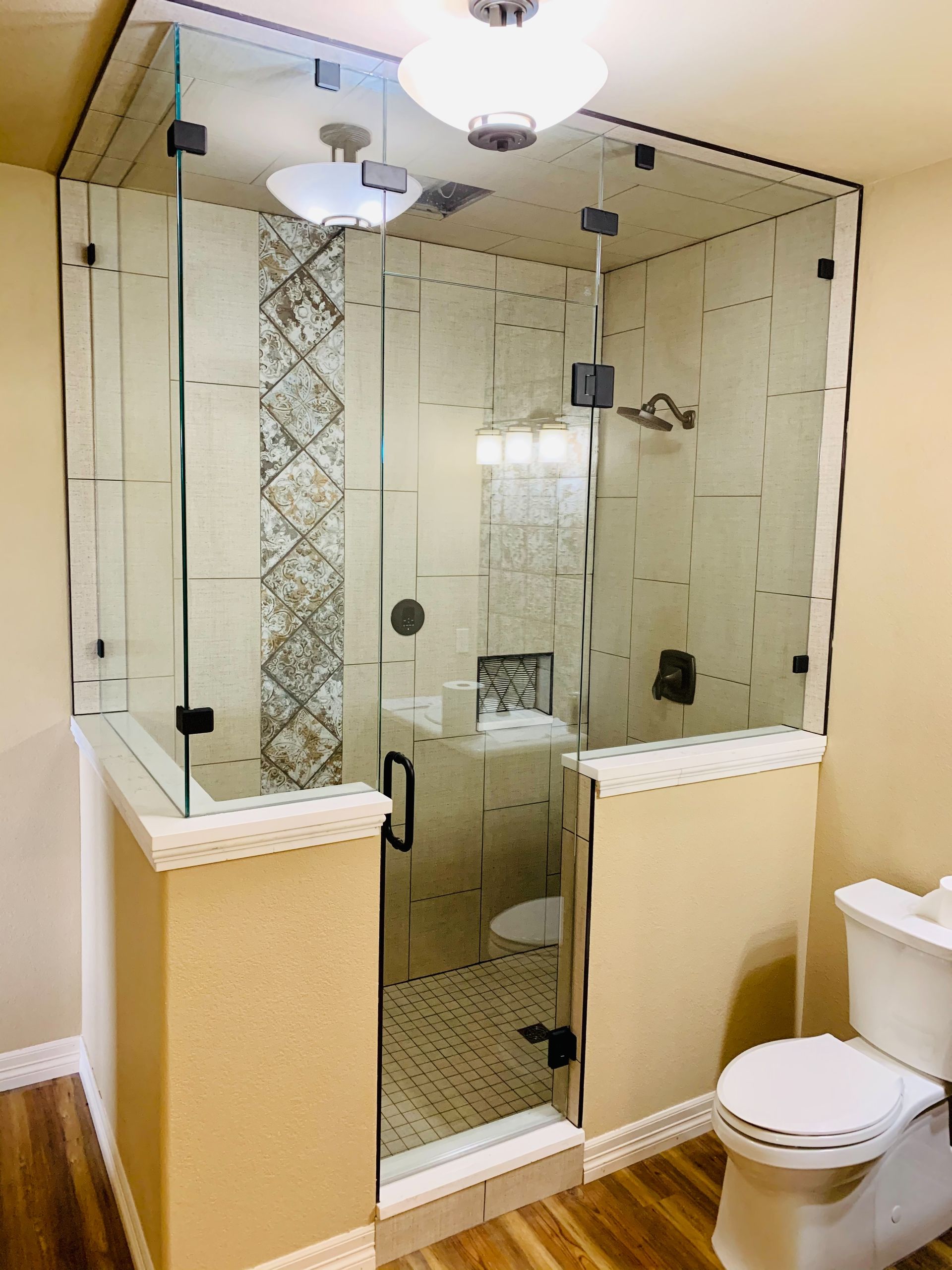 Glass-enclosed corner shower beside a white toilet in a beige bathroom with tiled walls and two lights