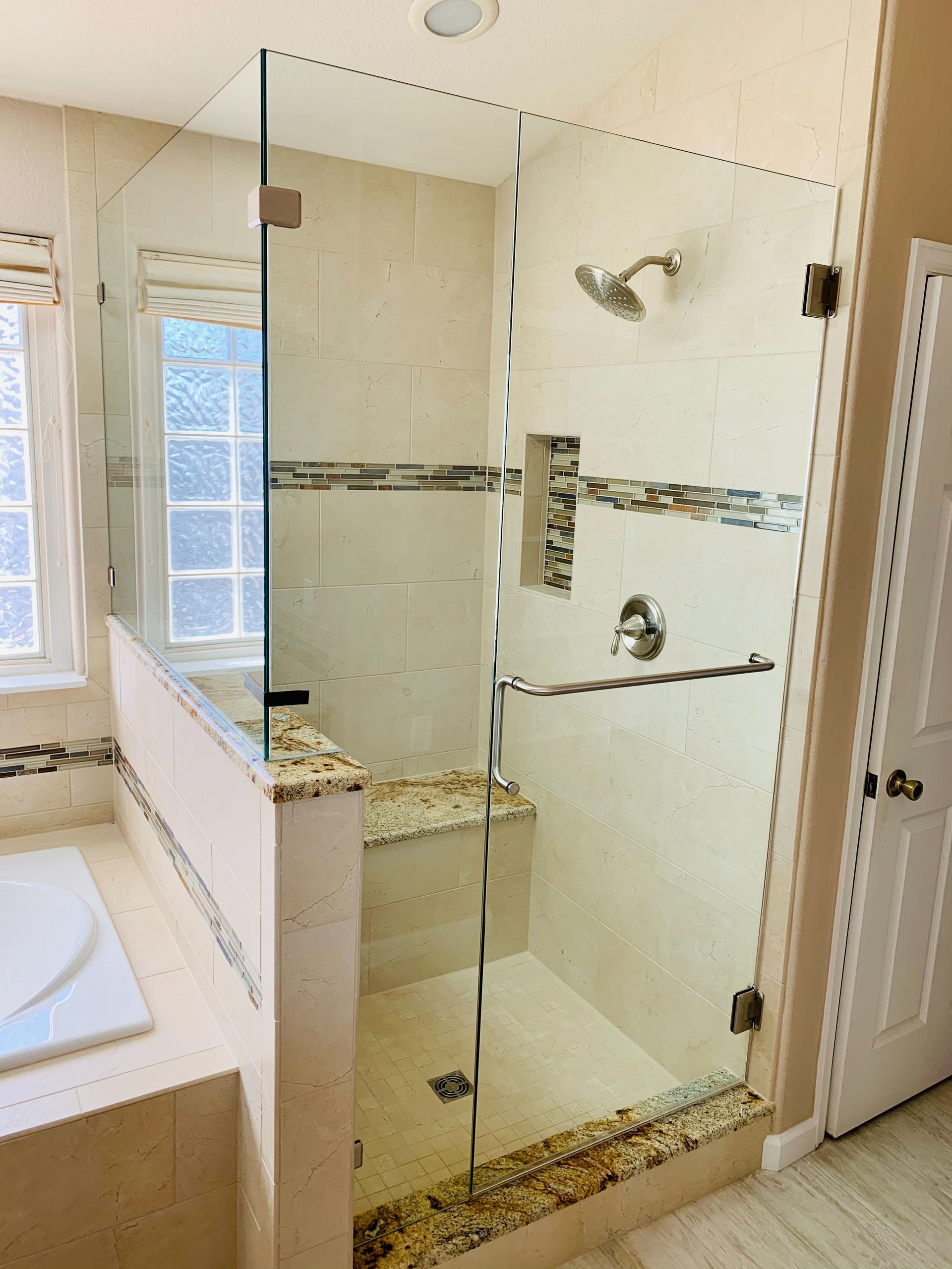 Glass-enclosed walk-in shower with beige tile, mosaic accent strip, and built-in bench beside a bathtub
