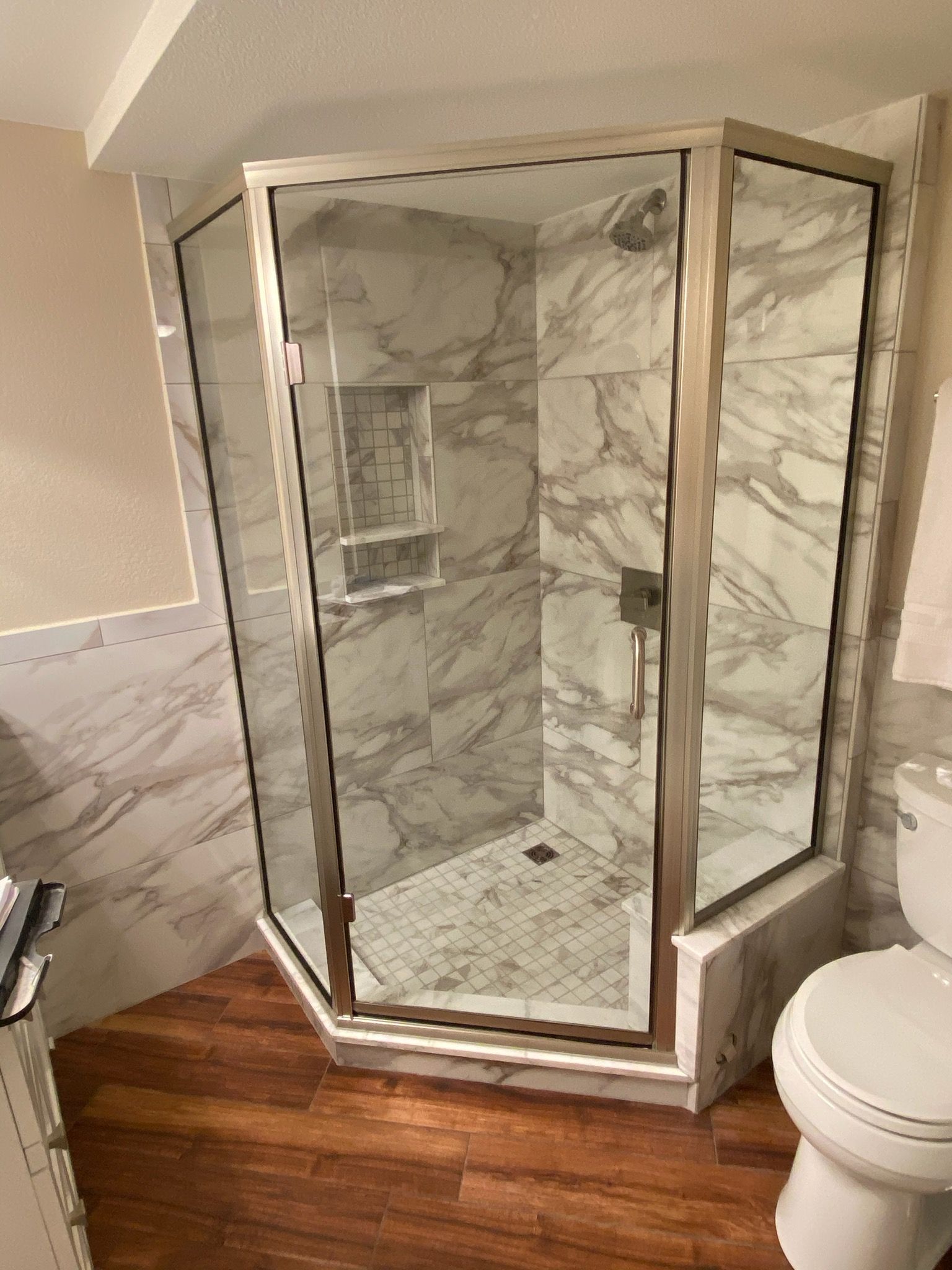 Corner glass shower enclosure with marble-look tile and wood bathroom floor
