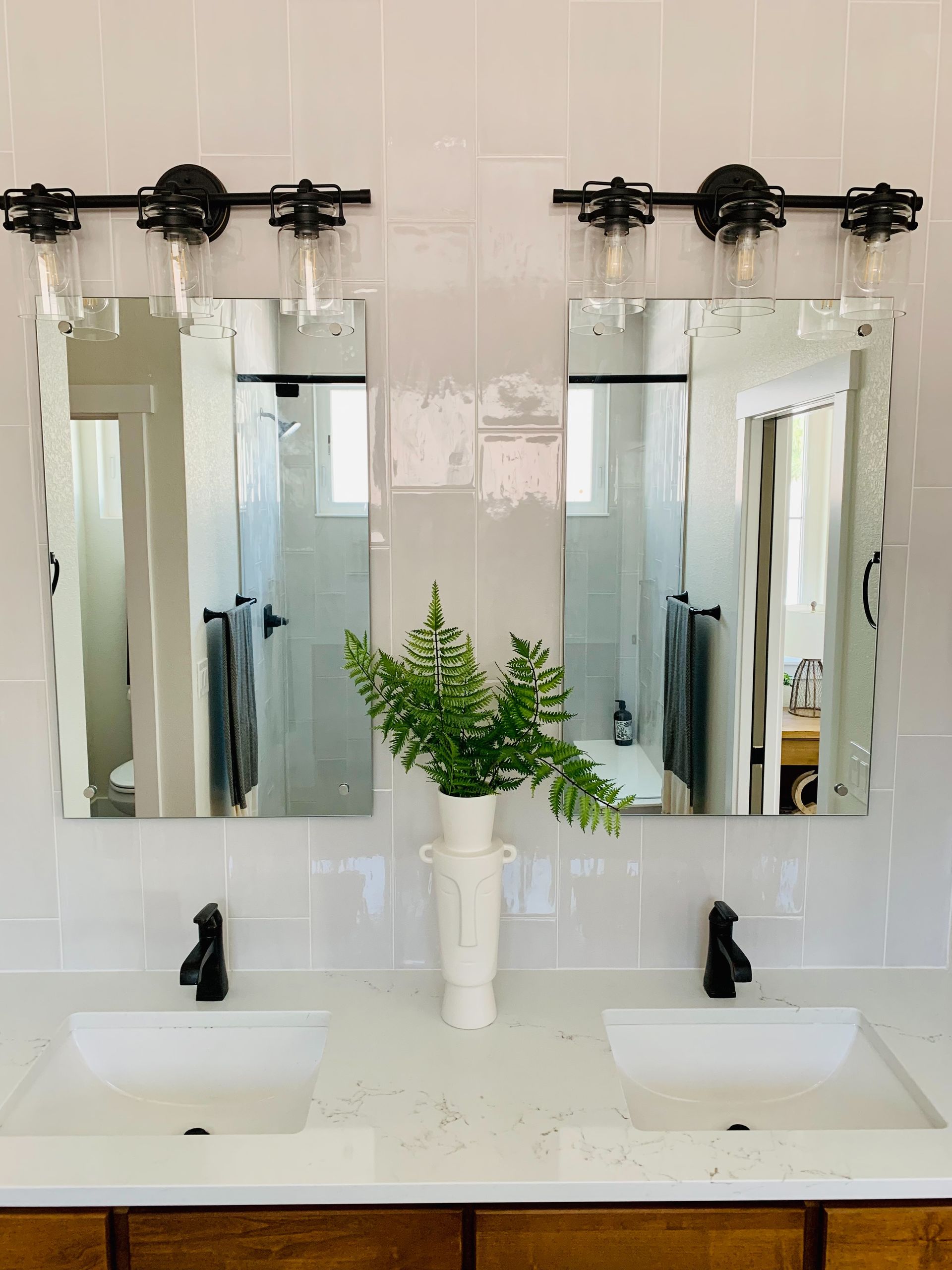 Double bathroom vanity with two mirrors, black faucets, and a vase of green foliage center.