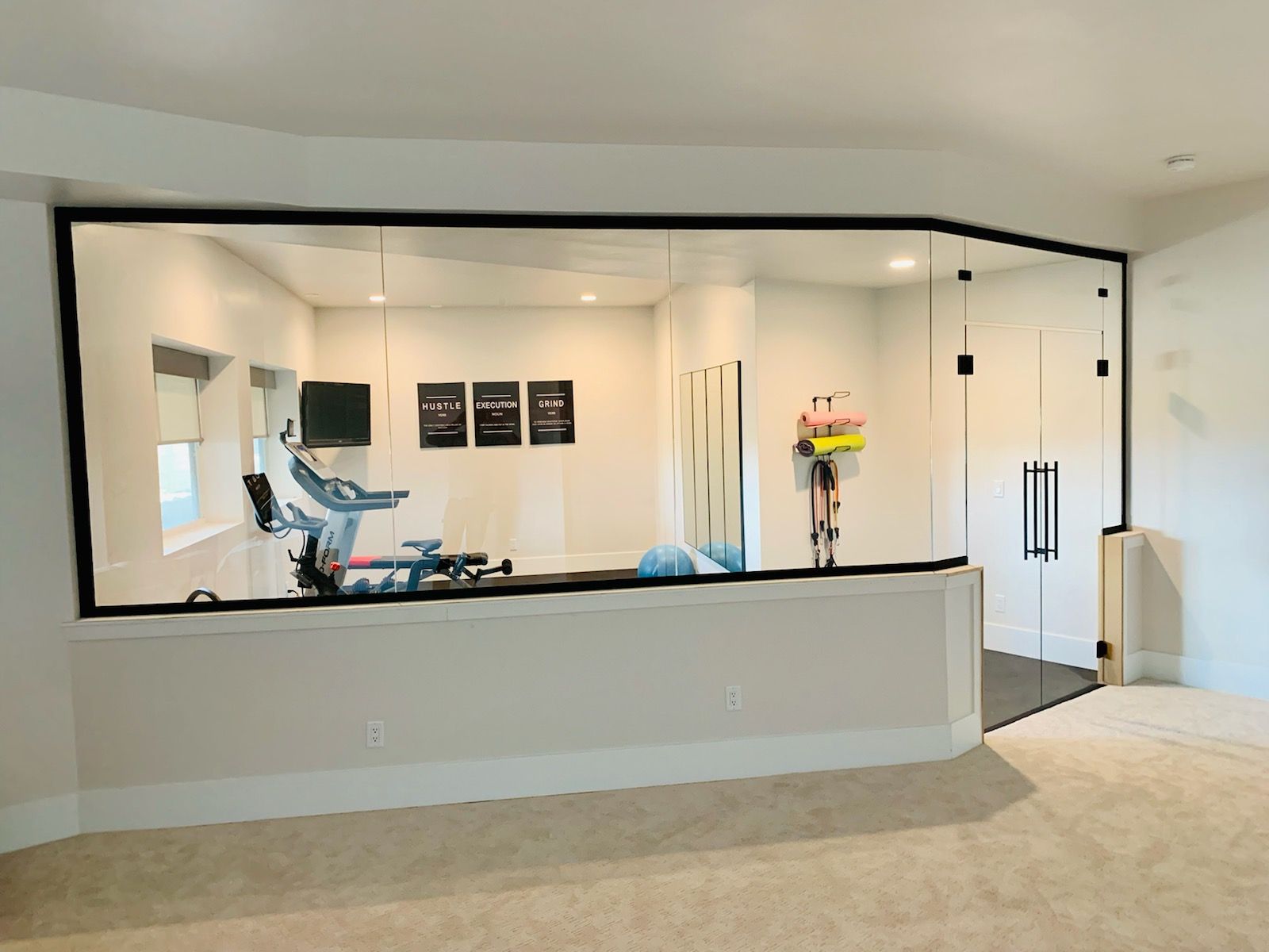 Modern home gym with exercise equipment, large mirror wall, and beige carpeted floor.