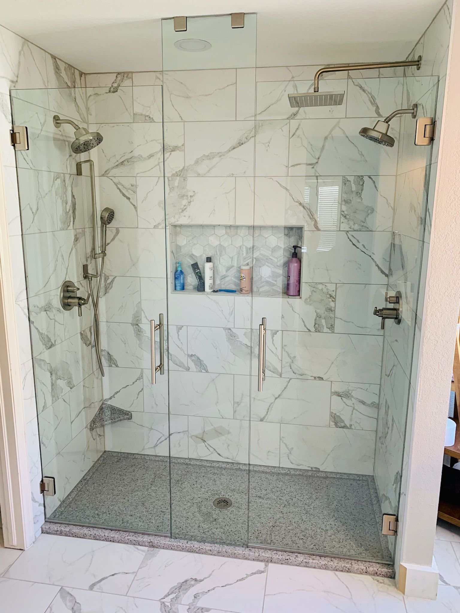 Walk-in shower with marble tile walls, dual showerheads, and a recessed shelf holding bottles.