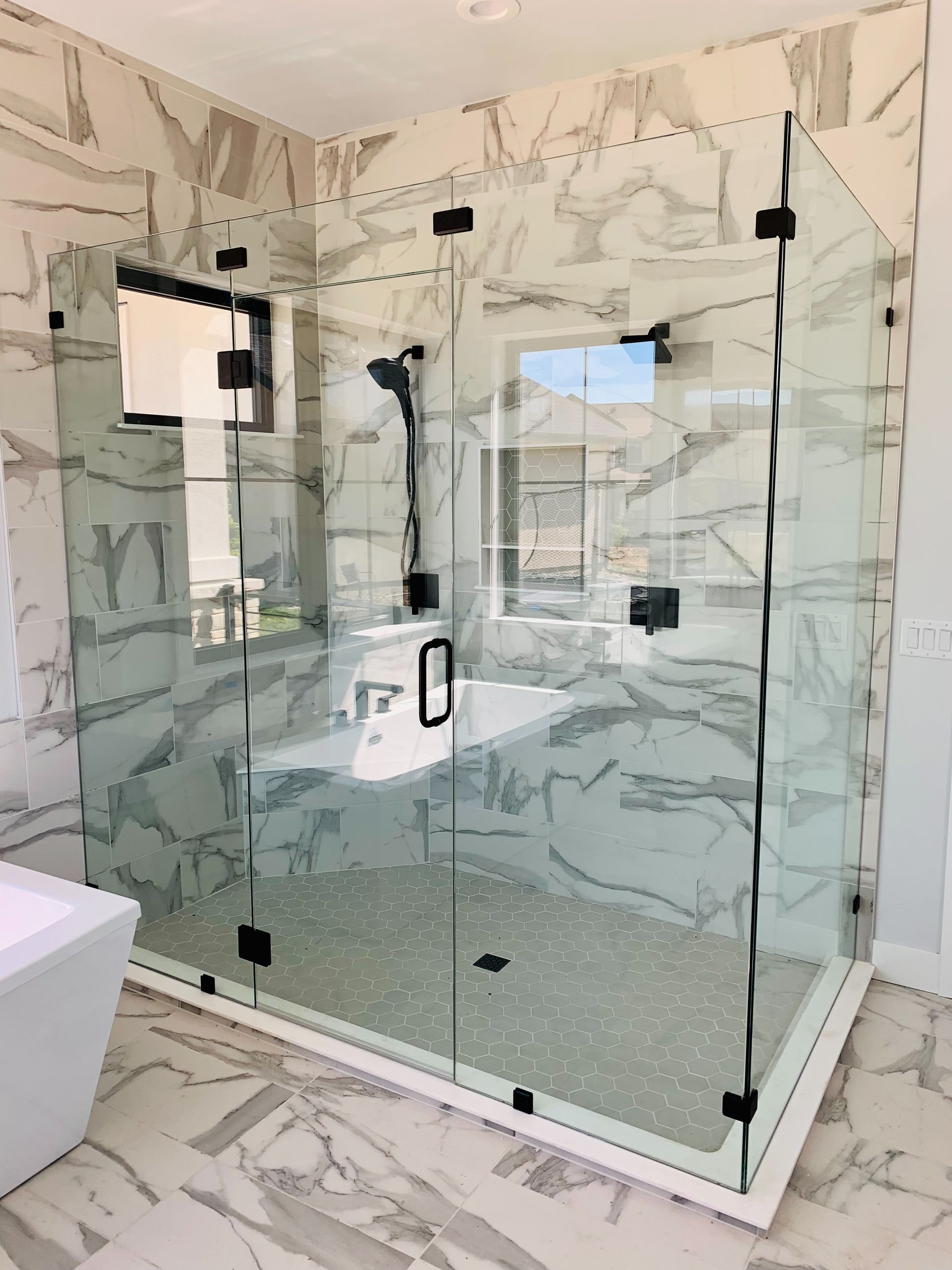 Modern bathroom with glass-enclosed marble shower, black hardware, and large window.