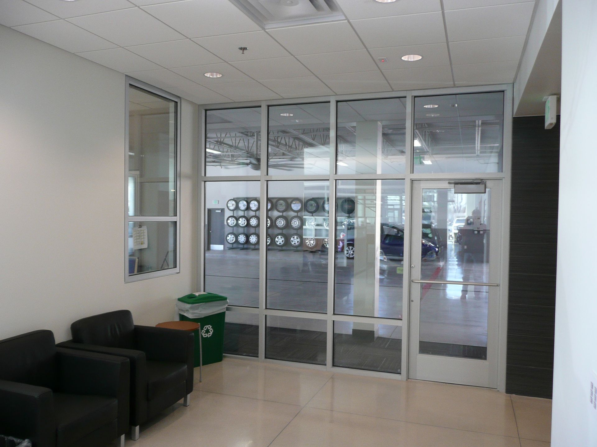Office lobby with black chairs, a green trash bin, and glass doors opening to a hallway.