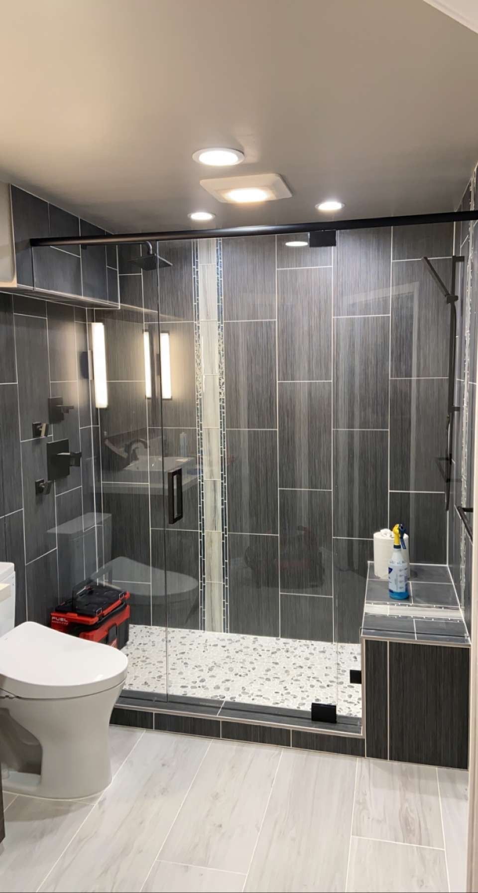 Modern bathroom with glass shower, dark tile walls, white floor, and vanity on the right