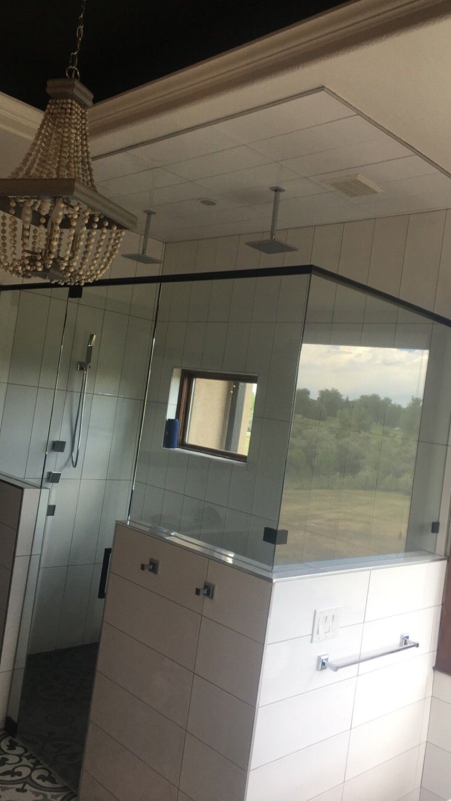 Dim bathroom with tiled shower, chandelier, wall mirror, and a window overlooking trees.
