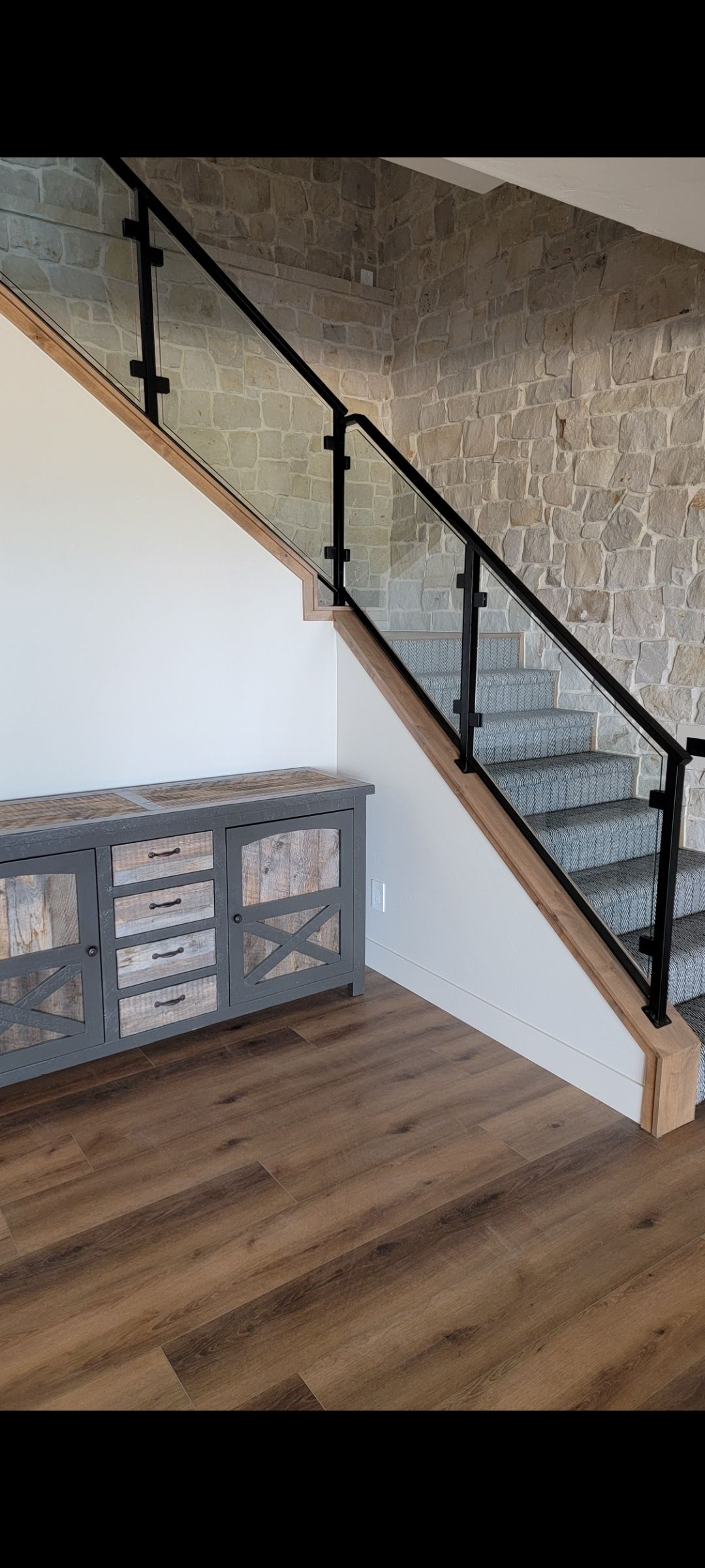 Staircase with black railing beside a stone wall in a room with wood flooring and a cabinet.