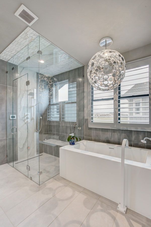Modern white bathroom with glass shower, freestanding tub, and pendant light, featuring gray tile and window blinds