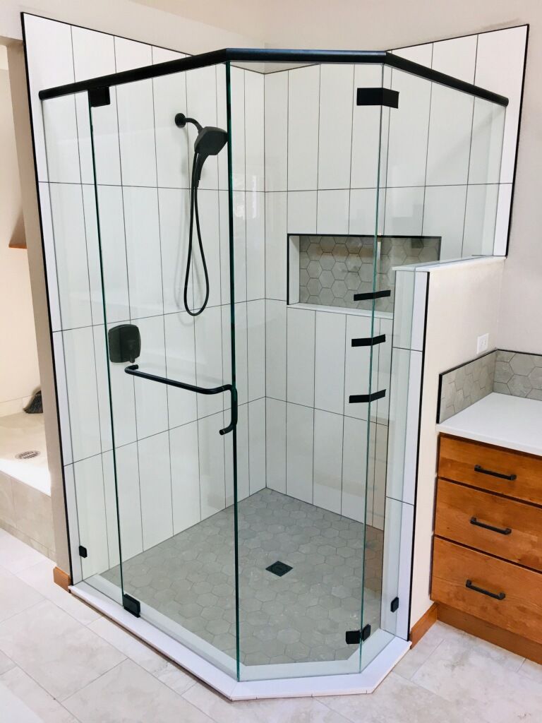 Glass corner shower with white tile walls, black hardware, a built-in shelf, and a small wood vanity nearby