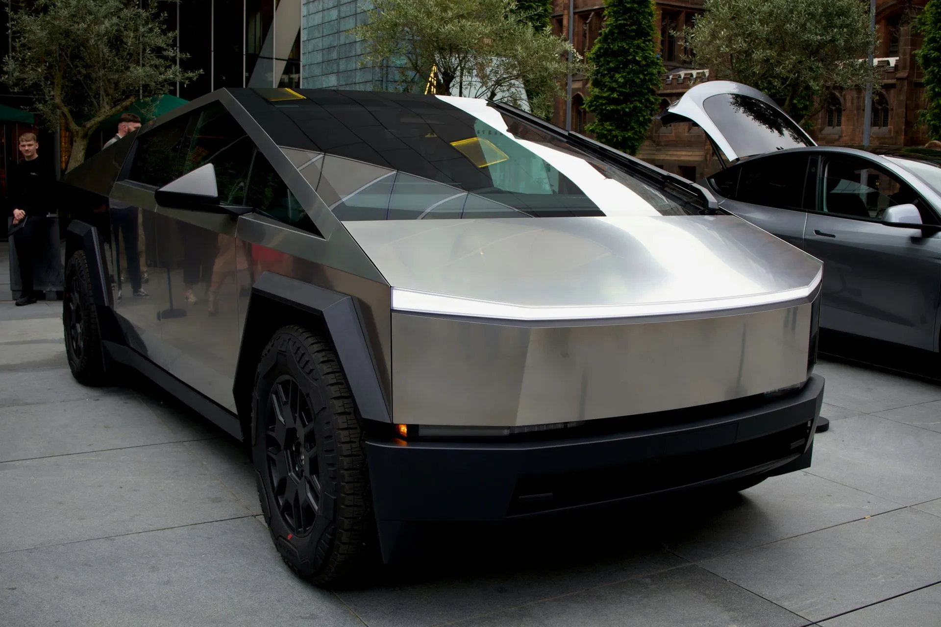 Silver Tesla Cybertruck parked on a street, with a black vehicle beside it and trees in the background