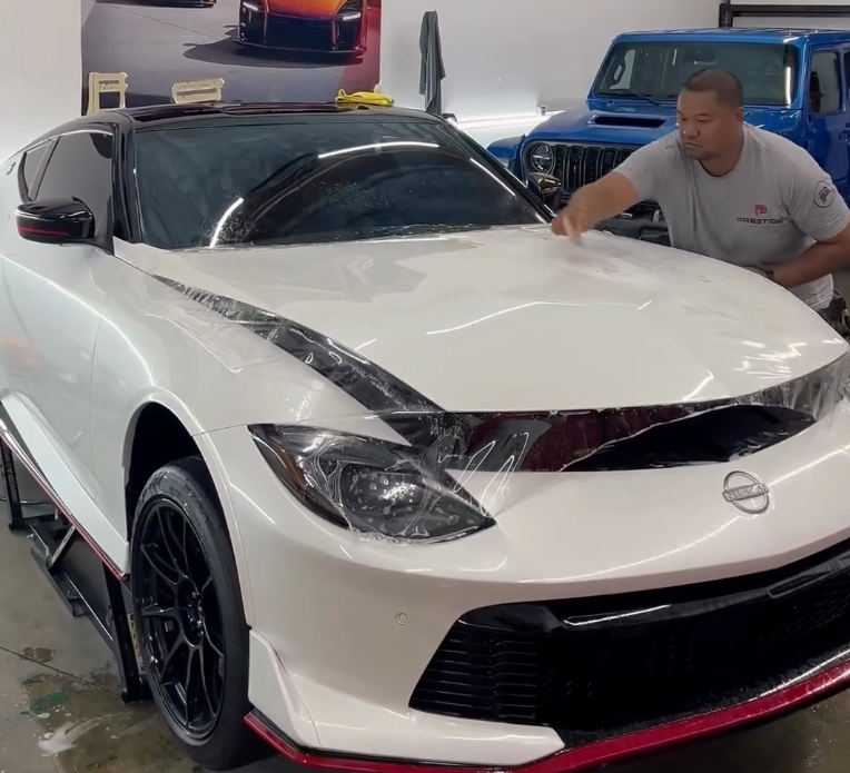 A man applies film to a white sports car hood in a shop. A blue Jeep sits behind it.