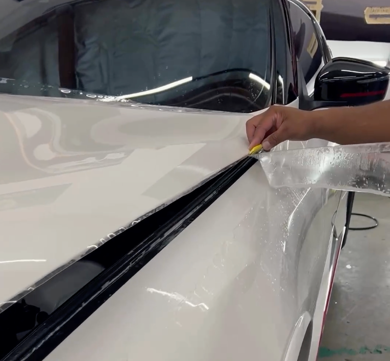 Applying protective film to a white car's hood. A hand uses a squeegee to smooth out the film.