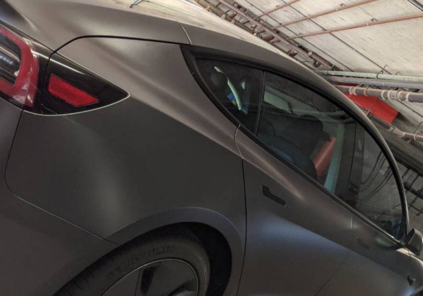 Gray Tesla Model Y, side view. The car is inside a building with visible pipes on the ceiling.