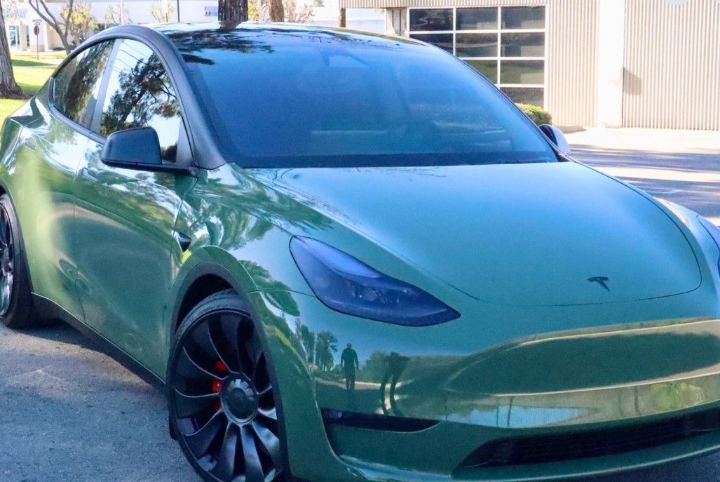 Green Tesla Model Y with black roof, wheels, and tinted windows parked outside.