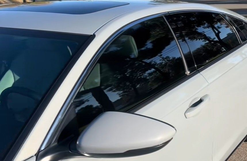 White car with tinted windows, side mirror, and sunroof reflecting trees.