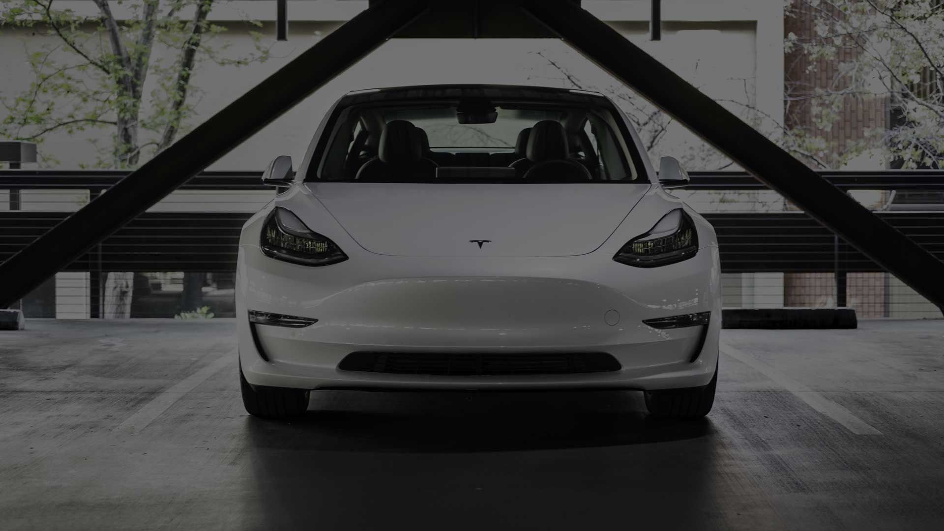 White Tesla parked indoors in a garage, front view with a geometric metal staircase behind it