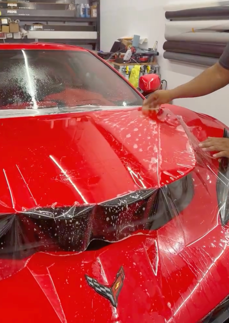 Applying protective film to a red Corvette's hood in a shop. Hands smoothing film with bubbles visible.