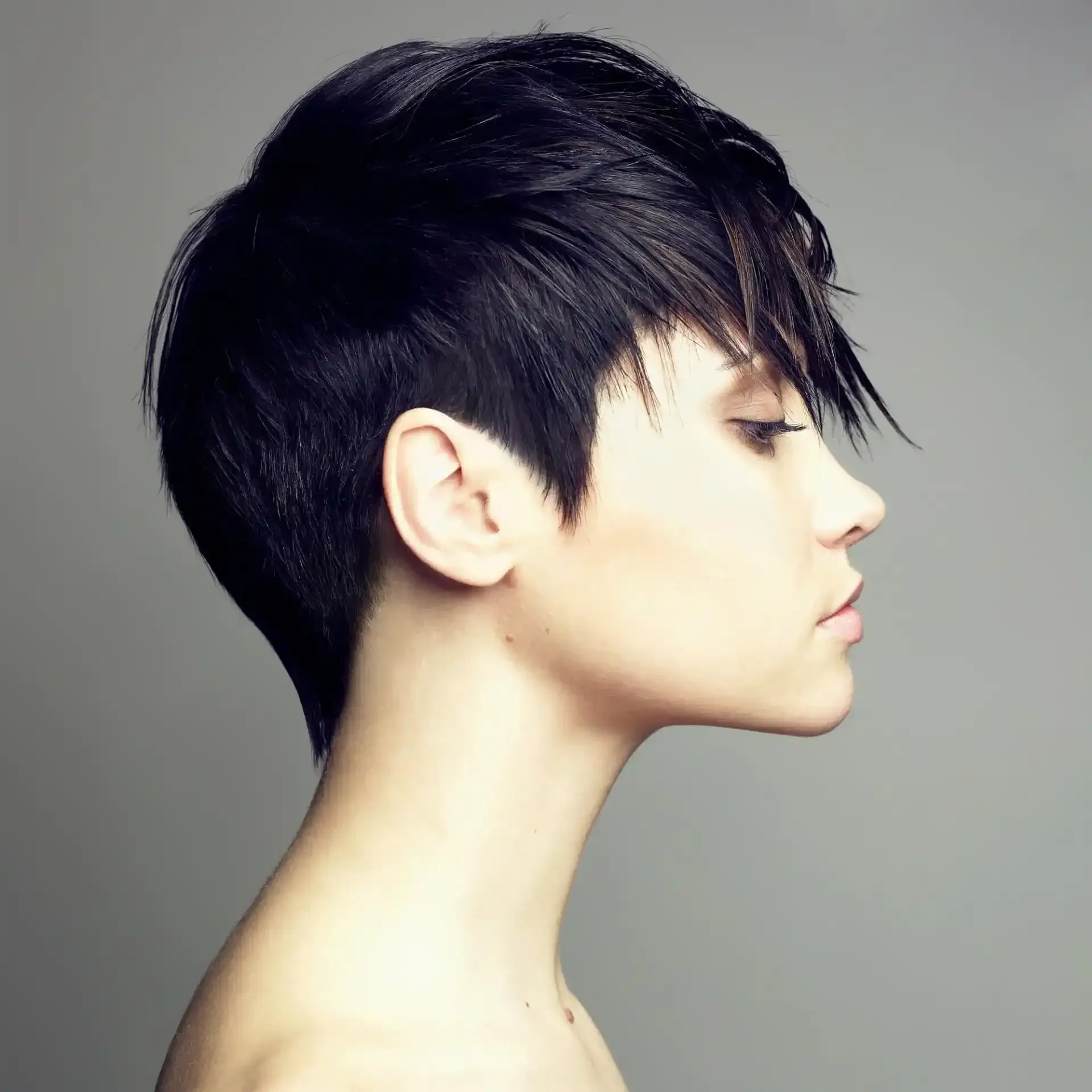 Donna con acconciatura corta e asimmetrica, capelli scuri, profilo laterale su sfondo grigio.