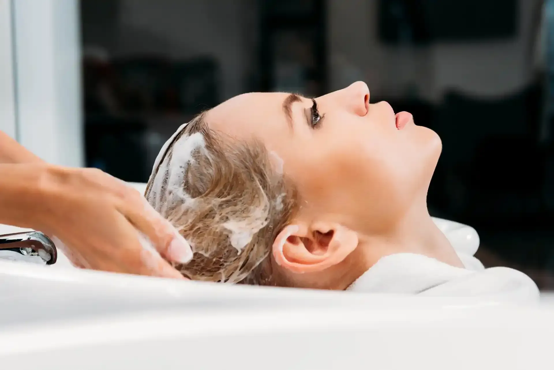 Donna sdraiata sul lavandino di un salone di bellezza, mentre viene lavata i capelli, si vede la schiuma bianca.