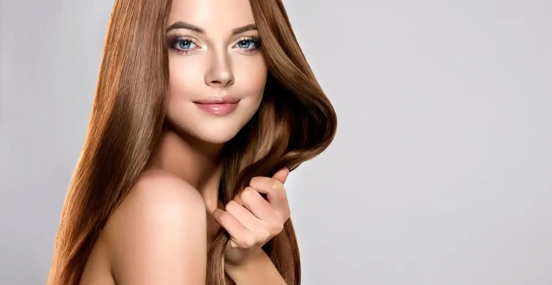 Donna con lunghi capelli castani lucidi, che tiene una ciocca e sorride, su sfondo grigio.