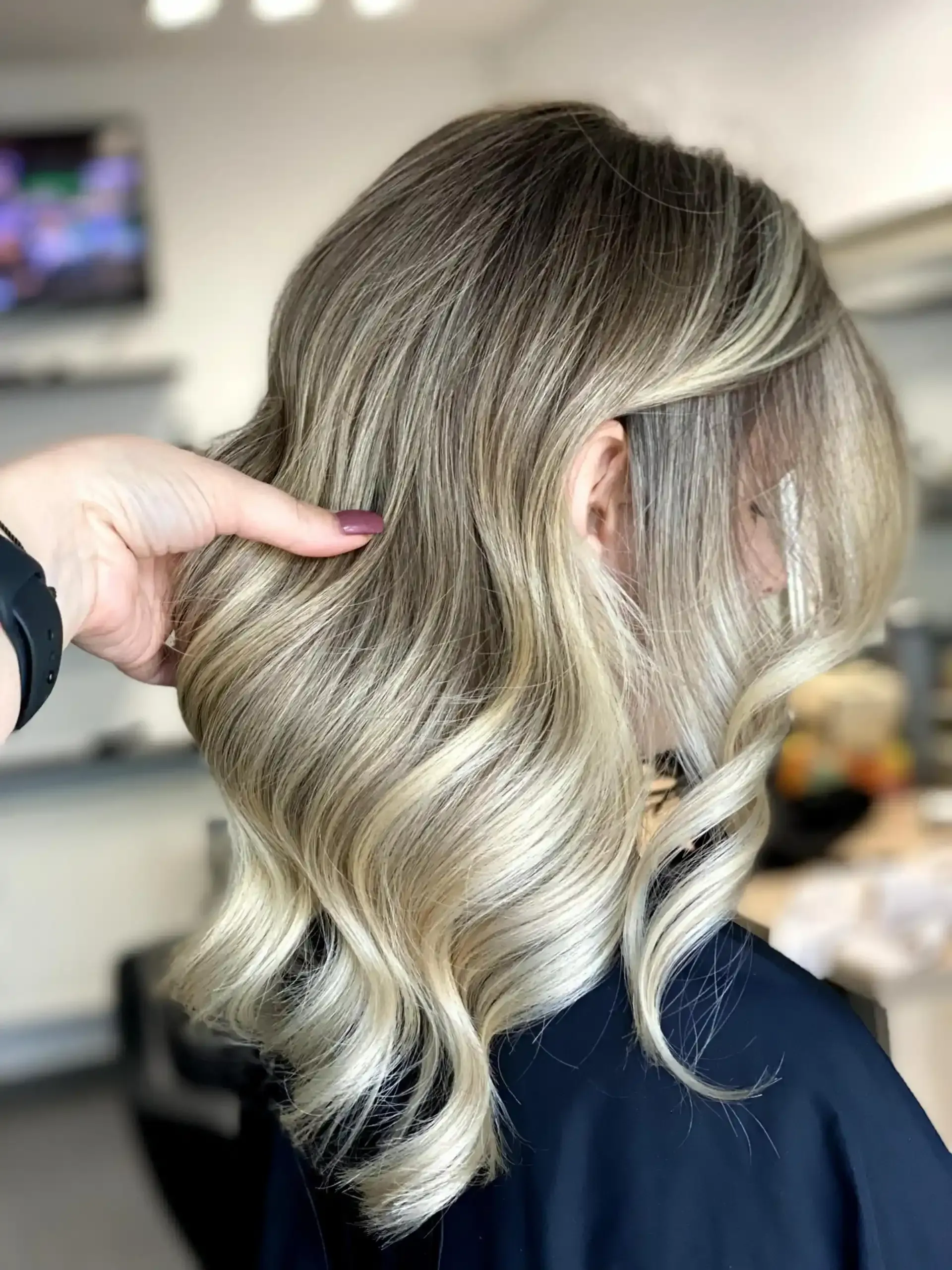 Capelli biondi ondulati e mèches di una donna, pettinati di lato e tenuti in mano; ambientazione da salone di bellezza.