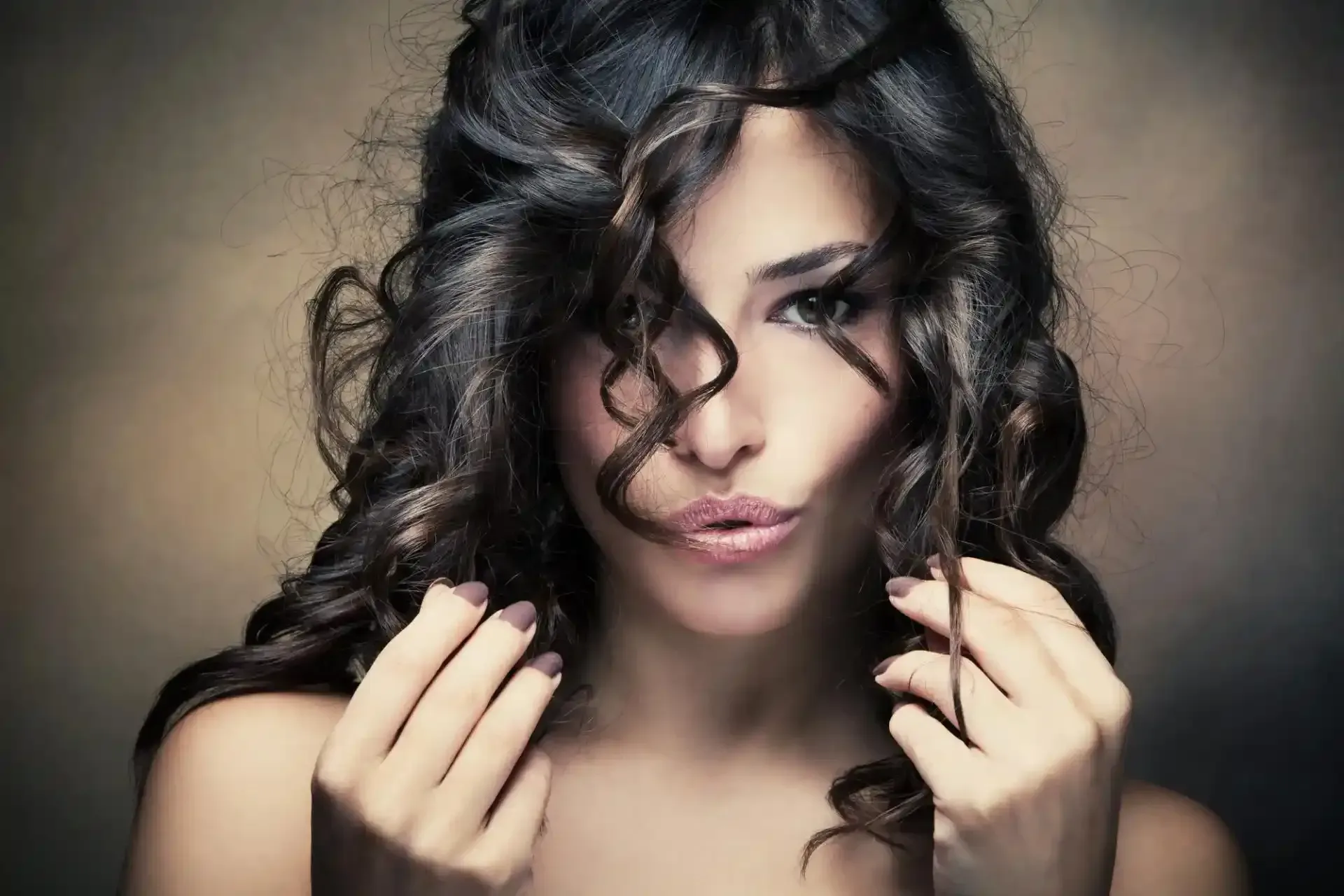 Donna con capelli scuri e ricci, mani vicino al viso, labbra serrate, su uno sfondo sfocato.