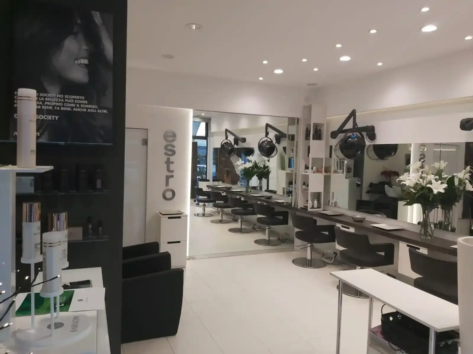 Interno di un salone da parrucchiere con postazioni per lo styling, specchi, piante e illuminazione intensa.