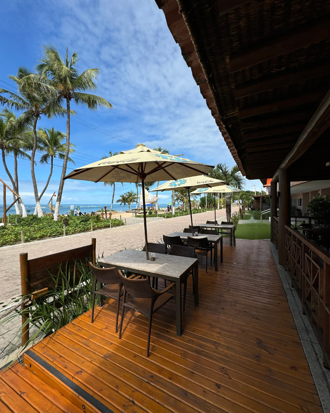 Restaurante ao ar livre com mesas, guarda-sóis e vista para a praia e as palmeiras. Dia ensolarado com céu azul.